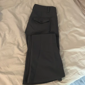 Mörkgrå kostymbyxor från Gina Tricot - Snygga mörkgrå kostymbyxor från Gina Tricot med två knappar i midjan, de är lågmidjade och är utsvängda i benen. Byxorna har en klassisk rak passform och en ficka med knapp bak. Perfekta för en clean och stilren look.