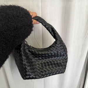 Svart flätad handväska i skinn - Snygg svart handväska med flätad design och kort handtag. Väskan är tillverkad i skinn och har en dragkedja upptill. Perfekt storlek för det viktigaste och ger en cool vibe till din outfit. bra skick!!💞
