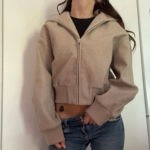 Beige croppad kofta  - Säljer en beige croppad kofta med dragkedja framtill och stor huva. Tröjan har breda muddar vid ärmslut och nederkant, samt långärmad passform. Storlek XS