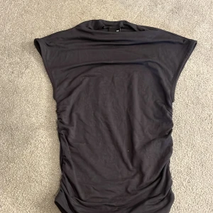 Topp från Gina Tricot XXS - Säljer en svart topp från Gina Tricot i storlek XXS. Toppen har en enkel och stilren design med rynkade sidor som ger en snygg siluett. Den är ärmlös och gjord i mjukt bomullsmaterial, perfekt för dig som gillar minimalistisk stil.