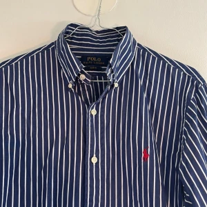 Randig blå skjorta Polo Ralph Lauren - Snygg blå skjorta från Polo Ralph Lauren i slim fit med vita ränder och klassisk krage. Långärmad modell med knappar framtill och ikonisk röd logobroderi på bröstet. Tillverkad i mjuk bomull.