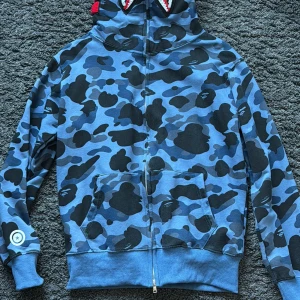 BAPE blå camo hoodie med shark-hood - Säljer en ikonisk BAPE hoodie i blått camo-mönster med svart och mörkblå detaljer. Tröjan har en fet shark-hood med broderade motiv och färgglada bokstäver, samt dragkedja hela vägen och känguruficka. Perfekt för dig som vill sticka ut med streetwear.