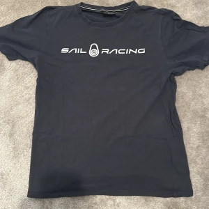 Mörkblå Sail Racing t-shirt - ✨ Sail Racing T-shirt ✨ Snygg mörkblå t-shirt från Sail Racing 🔵 med vit logotyp på bröstet och en mindre logga i nacken 🤍. 	•	👕 Klassisk rund hals och korta ärmar 	•	🧵 Tillverkad i mjuk bomull som känns skön mot huden 	•	🌟 Perfekt för dig som gillar stilren design och sportiga märken  En t-shirt som kombinerar komfort och sportig elegans. 
