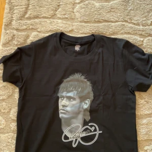 Svart t-shirt med tryck och signatur - Svart t-shirt i 100% bomull med rund hals och korta ärmar. På framsidan finns ett stort tryck av ett ansikte med cool frisyr och en vit signatur under. Baksidan är helt svart utan tryck. Perfekt för dig som gillar grafiska motiv.