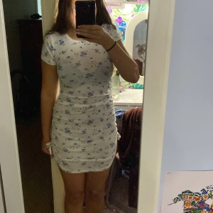 Vit blommig kortklänning från Shein - Supersöt vit kortklänning från Shein med blå blommor och snörning på sidorna. Klänningen har rund hals, korta ärmar och en tight passform som framhäver figuren. Perfekt för sommarens alla tillfällen, enkel att styla med sneakers eller sandaler.