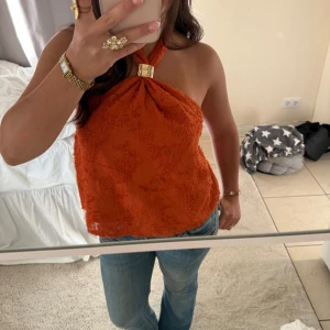 Orange omlott halterneck topp - Säljer en orange halterneck topp med omlott-detalj och snyggt guldfärgat spänne vid halsen. Toppen är ärmlös och har en lätt textur i tyget som ger en cool känsla. Perfekt till jeans och accessoarer för en trendig look.