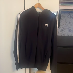 Svart Adidas hoodie med vita ränder - Svart hoodie från Adidas med klassiska vita ränder längs ärmarna och Adidas-logga på bröstet. Tröjan har huva, dragkedja framtill och långa ärmar. Perfekt för chill eller träning, snygg och sportig stil. Materialet är mjuk polyester.