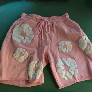 Rosa shorts från Denim Tears med snörning - Unika rosa shorts från Denim Tears med vita broderade blommor över hela plagget. Modellen har elastisk midja med snörning och är tillverkad i mjuk bomull. Perfekt för dig som vill sticka ut med coola detaljer och skön passform. Priset kan diskuteras via dm