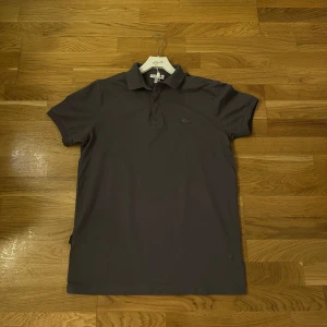 Mörkgrå Lacoste piké t-shirt slim fit - Snygg mörkgrå piké t-shirt från Lacoste i slim fit-modell. Klassisk krage och korta ärmar, med den ikoniska krokodilloggan ton-i-ton på bröstet. Tillverkad i mjuk bomull som känns skön mot huden. Perfekt för dig som gillar stilrena och enkla plagg.