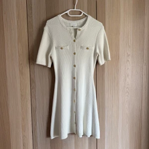 Vit stickad kortklänning från H&M - Supersöt vit stickad klänning från H&M med korta ärmar och rund hals. Klänningen har snygga guldfärgade knappar hela vägen framtill och två dekorativa bröstfickor. Ribbad struktur och lätt utställd nederdel ger en chill och feminin vibe.