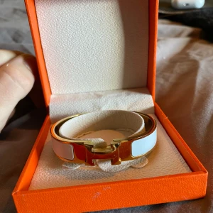  vit och guld armband - Snyggt  armband i vit och guld med klassisk H-detalj framtill. Armbandet har en stilren och modern design med blanka vita paneler och guldfärgad metall. Perfekt accessoar för dig som vill sticka ut med lyxig vibe och ikonisk look.