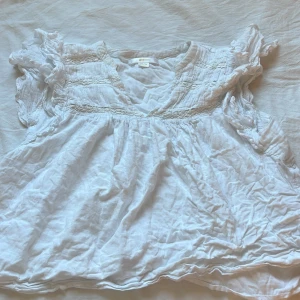 Vit blus med volang från Gina Tricot - Supersöt vit blus från Gina Tricot i luftig bomull. Blusen har v-ringad hals, korta volangärmar och smockdetaljer upptill. Perfekt för dig som gillar romantisk och chill stil. Passar dig som vill ha en lätt och flowy topp till sommaren. Har ett brunt märke vid armhålan men syns inte 