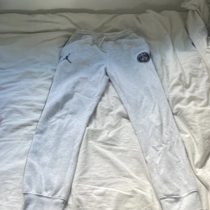 Grå PSG x Jordan mjukisbyxor XL - Snygga ljusgrå mjukisbyxor från Jordan x PSG med broderad logga på låret och blå ränder längs benen. Byxorna har snörning i midjan och muddar vid bensluten. Perfekta för chill eller sport. Materialet är mjuk bomullsmix.