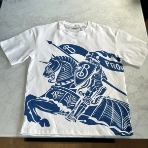 Vit Burberry t-shirt med blått tryck - Snygg vit t-shirt från Burberry med stort blått grafiskt tryck av riddare och flagga på framsidan. Klassisk rund hals och korta ärmar. Tillverkad i mjuk bomull som känns skön mot huden. Perfekt statement-plagg för dig som gillar exklusiva märken och cool design.