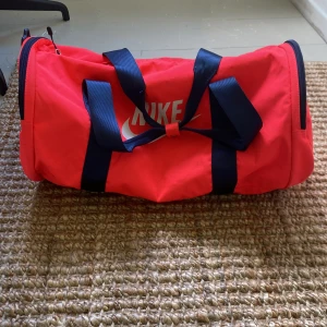 Neon rosa träningsväska från Nike - Säljer en neon rosa träningsväska från Nike med marinblå detaljer och handtag. Väskan har cylinderform, flera dragkedjor och ett stort fack samt mindre fack på sidan. Perfekt för gymmet eller sportaktiviteter. Nike-logga i vitt på framsidan.