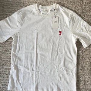 Vit t-shirt från Ami med röd brodyr - Vit t-shirt från Ami Alexandre Mattiussi i mjuk bomull. Klassisk rund hals och korta ärmar. På bröstet finns Ami:s ikoniska broderade logga med rött hjärta och bokstaven A. Enkel och clean design som känns fräsch och modern.