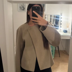 Beige omlott kappa i ullmix - Snygg beige kappa med omlottknäppning och en stor knapp vid halsen. Jackan har en stilren och minimalistisk design med raka linjer och lång ärm. Perfekt för dig som gillar en clean och modern look. Materialet känns mjukt och har en lyxig känsla.