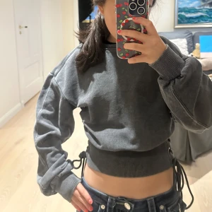 Mörkgrå croppad sweatshirt med snörning - Säljer en mörkgrå croppad sweatshirt med snygg snörning i sidorna och breda muddar vid ärmslut och midja. Tröjan har lång ärm och en loose passform som ger en chill vibe. Perfekt för dig som vill ha något unikt i garderoben. Använd en gång!