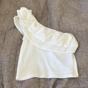 Vit offshoulder topp med volang - Superfin vit topp med offshoulder-design och bred volang med broderade hålmönster. Sitter snyggt med en axel bar och har en lätt ribbad struktur i tyget. Perfekt för dig som gillar romantiska detaljer och somrig stil.