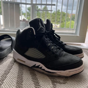 Nike Air Jordan 5 Moonlight - Säljer ett par svarta Nike Air Jordan 5 sneakers med högt skaft och klassisk Jumpman-logga på både plös och häl. Skorna har snörning, vit och svart sula med genomskinliga detaljer och meshpaneler på sidorna. Perfekt för dig som gillar streetstyle och ikoniska basketskor.