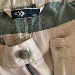 Beige shorts från 157, storlek L - Snygga beige shorts från 157 med grå detaljer vid fickor och dragkedjor. Modellen är midwaist och har en tight passform. Materialet känns som bomull med inslag av syntet, vilket gör dem mjuka och stretchiga. Perfekta för sommarens äventyr och har praktiska fickor med dragkedja.