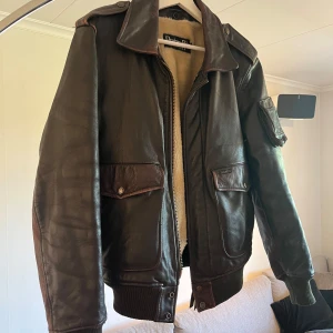 90-tals vintage A2 Pilot jacka i skinn. - Riktigt fet mörkbrun skinnjacka från Petroff med schysst fuskpälsfoder. Klassisk bomberstil med stora fickor, axelklaffar och ribbade muddar. Jackan har dragkedja och coola detaljer i läder. Perfekt för dig som vill sticka ut med vintagevibbar.