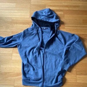 Blå hoodie från Dressmann med dragkedja - Blå hoodie från Dressmann i storlek M. Tröjan har huva med snörning, hel dragkedja och två fickor fram. Materialet är mjukt och känns som bomull, med diskreta vågiga linjer i mönstret. Perfekt för chill dagar eller när du vill vara bekväm.