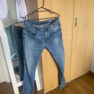Blå jeans från Replay - Säljer ett par snygga blå jeans från Replay med klassisk femficksdesign och coola slitningar. Jeansen har normal passform och är tillverkade i mjukt denimtyg med Replay-logga på fickan och knappen. Perfekta för en avslappnad och trendig look.