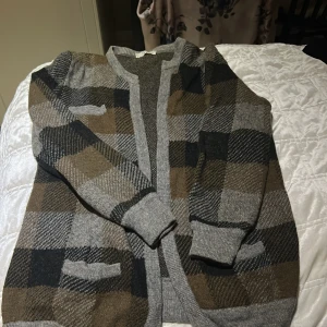 Rutig kofta från Valentino Studio - Säljer en snygg rutig kofta från Valentino Studio i grått, brunt och svart. Koftan har långa ärmar, ribbade muddar och två fickor framtill. Materialet är mjukt och stickat, perfekt för dig som gillar klassisk stil med en modern twist.