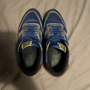 Valentino Rockrunner blå sneakers - Snygga Valentino Rockrunner sneakers i blått med grå mockadetaljer och bruna accenter vid hälen. Skorna har snörning, platt sula och rund tå. Ovandelen är i mesh och mocka, med coola nitar på hälen som ger extra edge. Perfekt för dig som gillar exklusiva sneakers med streetkänsla.