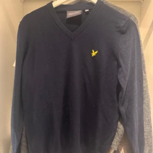 Marinblå stickad tröja Lyle & Scott - Snygg marinblå stickad tröja från Lyle & Scott med gul broderad logga på bröstet. V-ringad hals och långärmad modell som passar perfekt till jeans eller chinos. Mjuk och skön kvalitet som håller värmen under kyliga dagar.