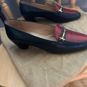 Celine loafers med klack, marinblå & röd - Snygga loafers från Celine Paris i marinblått och rött skinn med guldfärgade metalldetaljer över vristen. Skorna har en låg klack och rund tå, vilket ger en klassisk och stilren look. Perfekta för dig som gillar att sticka ut med exklusiva detaljer.