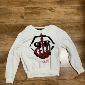 Vit sweatshirt från Philipp Plein - Säljer en vit sweatshirt från Philipp Plein med ett stort tryck på framsidan i svart och rött, där ett skelett visar långfingret och texten 'fuck off!' är broderad. Tröjan har ribbade muddar och rund hals. Perfekt för dig som gillar edgy streetwear.