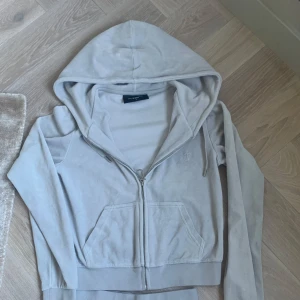 Ljusgrå hoodie från Juicy Couture - Säljer en ljusgrå hoodie från Juicy Couture i mjuk velour med dragkedja och huva. Tröjan har långa ärmar, fickor fram och ett broderat JC-monogram på bröstet. Perfekt för chill dagar och har en klassisk, clean look. RIKTIG