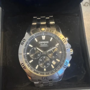 Champion Roadster kronografklocka - Snygg och sportig Champion Roadster kronografklocka i silverfärgat rostfritt stål. Klockan har svart urtavla med flera små urtavlor, tydliga index och datumvisning. Robust länkarmband och vridbar bezel ger en cool och klassisk look. Fungerar utan konstigheter men är i behov av batteribyte. 