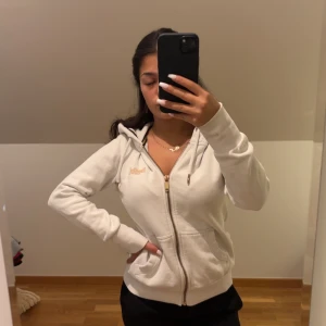 Vit hoodie från Superdry med dragkedja - Säljer en vit hoodie från Superdry med guldfärgade detaljer och broderad logga på bröstet. Tröjan har huva med snörning, två fickor fram och dragkedja. Mjuk bomullskänsla och ribbade muddar vid ärmslut och nederkant. Perfekt för kalla dagar då den är väldigt varm.