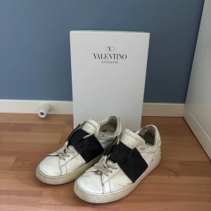 Valentino Garavani Open sneakers vit/svart - Säljer ett par Valentino Garavani Open sneakers i vitt skinn med bred svart läderdetalj över mitten. Klassisk rund tå, och snörning. Snygg och stilren design som sticker ut med den kontrasterande svarta panelen. Box medföljer med allt på bilderna. Slitningar undertill på sulan se bild nr 4. PRISET ÄR EJ FAST. 