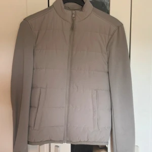 Beige quiltad pufferjacka med dragkedja - Snygg beige pufferjacka med quiltad framsida och hel dragkedja. Jackan har en stilren, rak passform och högt krage. Perfekt för dig som gillar minimalistisk stil och vill ha en jacka som funkar till många outfits.