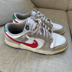 Nike Dunk Low  - Nike Dunk Low sneakers med vit och beige ovandel i skinn och mocka, röd swoosh på sidan och gröna detaljer vid hälen. Yttersulan är blå och mellansulan har en gul ton. Perforerad tåbox och snörning framtill. Liten gul swoosh på tån för extra stilpoäng.