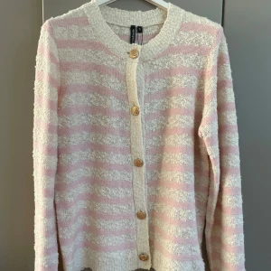 Randig kofta från Soulmate i rosa och gräddvitt - Säljer en söt kofta från Soulmate med ljusrosa och vita ränder. Koftan har lång ärm, rund hals och snygga guldfärgade knappar framtill. Materialet är stickat med en fluffig struktur som ger extra mysig känsla. Perfekt för dig som gillar pastell och mjuka vibes.