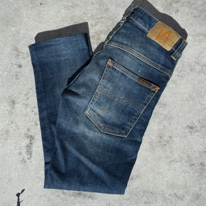 Mörkblå jeans från Nudie Jeans - Snygga mörkblå jeans från Nudie Jeans med klassisk femficksdesign och kontrastsömmar. Jeansen har en rak passform och är tillverkade i slitstarkt denim med coola detaljer som Nudie-loggan på läderpatchen bak. Perfekta för dig som gillar stilrena och tidlösa jeans. Av klippta och sydda som ni ser på bild 4! Passar dig perfekt om du är ungefär 155!