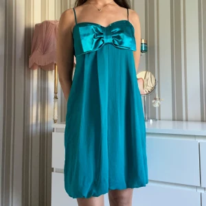 Turkos kortklänning med rosett från gina  - Supersöt turkos kortklänning med tunna axelband som går att justera och stor rosett framtill i glansigt tyg. 