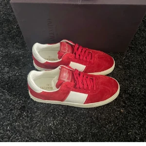 Valentino flycrew röda  - Snygga röda och vita sneakers från Valentino Garavani med mocka på ovandelen och vita läderdetaljer. Skorna har snörning, rund tå och cool kamouflagemönstrad sula. Perfekt för dig som vill sticka ut med exklusiva och trendiga sneakers.