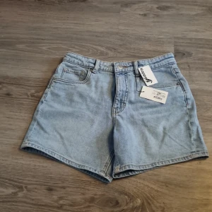 Ljusblå jeansshorts från Kappahl - Snygga ljusblå jeansshorts från Kappahl i klassisk femficksmodell. De har hög midja, raka ben och coola bakfickor med knappdetalj. Perfekt för dig som gillar en chill och enkel stil på sommaren. Passar till allt och är superbekväma med mjukt denimtyg.