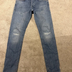 Blå skinny jeans storlek 32/34 - Säljer ett par blå skinny jeans i storlek 32/34 med slitna detaljer på knäna. Jeansen har klassisk femficksdesign och är tillverkade i mjukt denimtyg som sitter tajt längs benen. Perfekt för dig som gillar en smal siluett och lite sliten look.