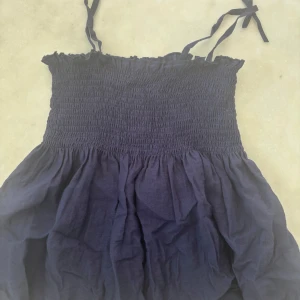 Marinblå kort klänning med smock - Säljer en marinblå topp  med smockad överdel och tunna axelband med russeter. Kjoldelen är luftig och har ett skirt fall. Perfekt för varma dagar och enkel att styla med sneakers eller sandaler.