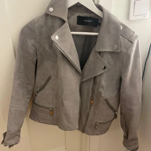 Grå mockajacka från Vero Moda - Snygg grå mockajacka från Vero Moda med cool bikerstil. Jackan har flera silvriga dragkedjor, bred krage med tryckknappar och detaljer på axlarna. Materialet är mjuk mocka och passformen är normal. Perfekt för dig som vill ha en stilren och edgy look.