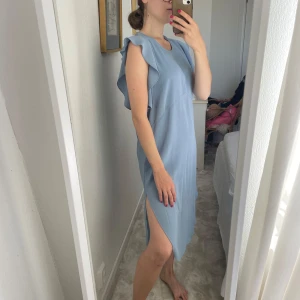 Ljusblå omlottklänning med volang - Säljer en ljusblå klänning med volangdetaljer vid axlarna och rund halsringning. Klänningen är ärmlös, har en omlottliknande skärning och en slits på sidan som ger en chill vibe. Materialet känns lätt och flowy, perfekt för varma dagar.