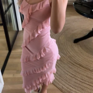 Rosa volangklänning med smala axelband - Säljer en superfin rosa klänning med smala axelband och flera lager volanger som ger en riktigt snygg och lekfull look. Klänningen är figurnära och har en kort längd, perfekt för dig som gillar att sticka ut. Materialet känns mjukt och följsamt mot kroppen.