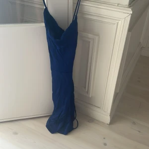 Blå långklänning från Zara - Säljer en snygg blå långklänning från Zara i storlek S. Klänningen har smala axelband, omlott-detalj i ryggen och en elegant, figurnära passform. Materialet är mjukt och har ett lätt glansigt utseende som ger en lyxig känsla. Perfekt för dig som gillar stilrena och trendiga plagg.